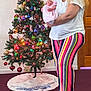 woman, baby, christmas_tree, holiday, ornaments, lights, indoor, carpet, striped_pants, white_tshirt, glasses, tattoo, door, garland, festive, smile, holding, peaceful, decorations, celebration