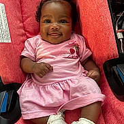 Malani is registered to the contest to win money with this photo: baby, infant, smile, pink_clothing, red_seat, cushion, cute, child, happy, person, portrait, sitting, indoors, sock, curly_hair, face, skin, hand, footwear, car_seat