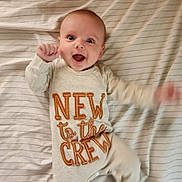 Dawsyn is registered to the contest to win money with this photo: baby, infant, smiling, lying_down, bed, striped_bedspread, clothing, long_sleeve, onesie, text_on_clothing, happy, cute, cheerful, hand_motion, face, head, eyes, mouth_open, skin, indoors