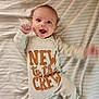 baby, infant, smiling, lying_down, bed, striped_bedspread, clothing, long_sleeve, onesie, text_on_clothing, happy, cute, cheerful, hand_motion, face, head, eyes, mouth_open, skin, indoors