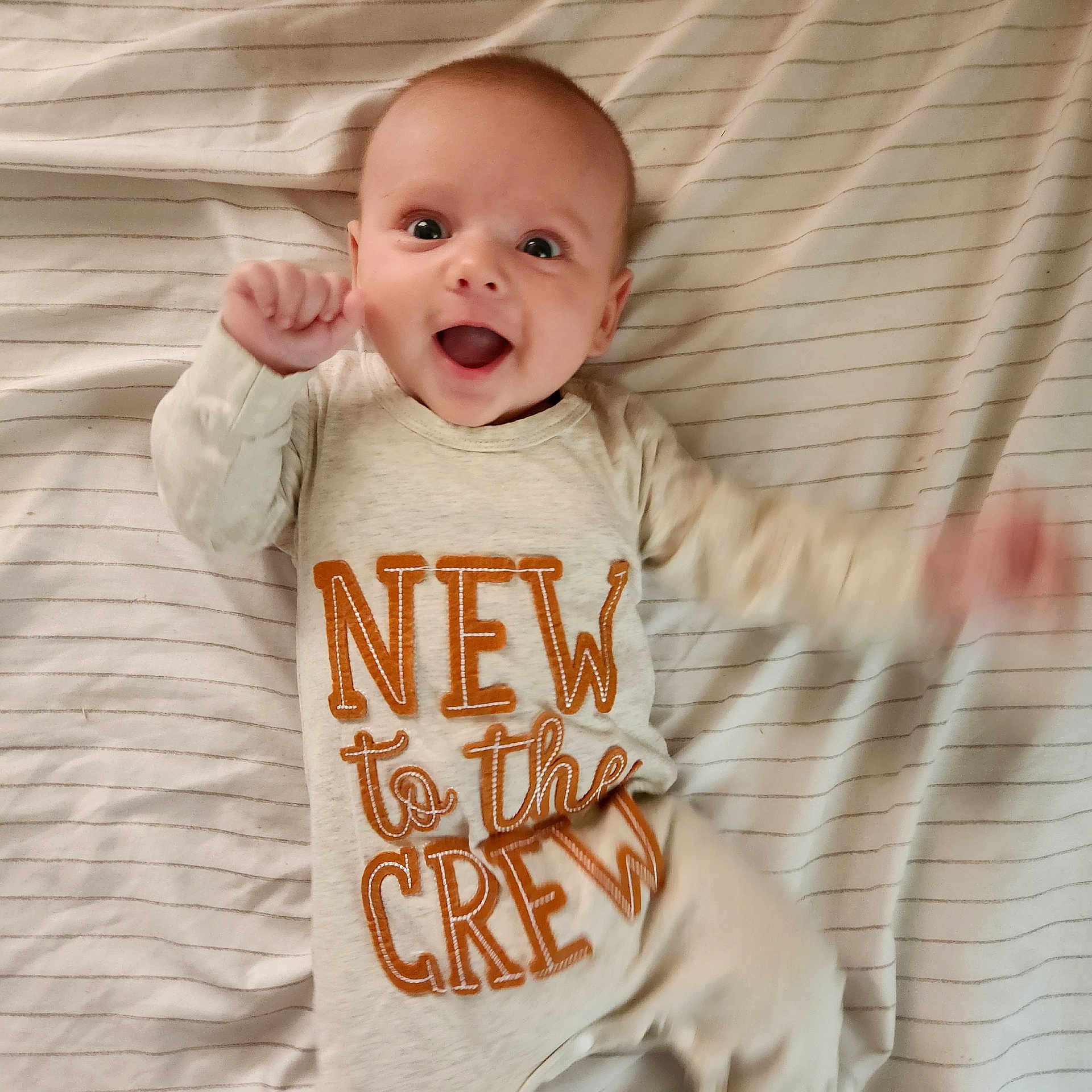 Dawsyn is registered to the contest to win money with this photo: baby, infant, smiling, lying_down, bed, striped_bedspread, clothing, long_sleeve, onesie, text_on_clothing, happy, cute, cheerful, hand_motion, face, head, eyes, mouth_open, skin, indoors