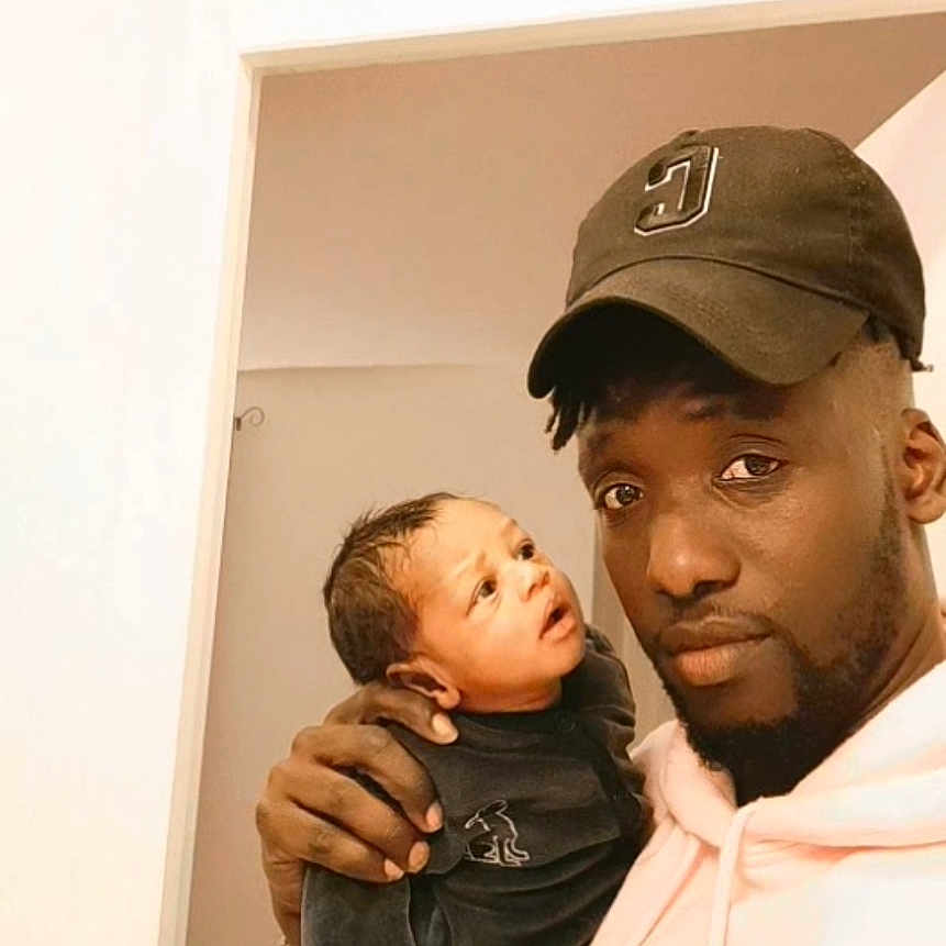 Bakary Diop a rejoint le concours — aidez-le/la à gagner de superbes lots ! adult, baby, bed, bedroom, cap, casual, child, clothing, curious, expression, face, hand, holding, home, hoodie, indoor, people, person, portrait, skin