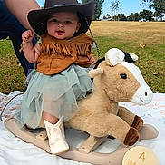 Lucy joined the competition — help win amazing prizes! baby, blanket, blue_sky, child, cowboy_hat, cute, fringed_vest, grass, happy, holding_hand, infant, milestone_sign, outdoor, park, person, playful, plush_toy, rocking_horse, smiling, white_boots