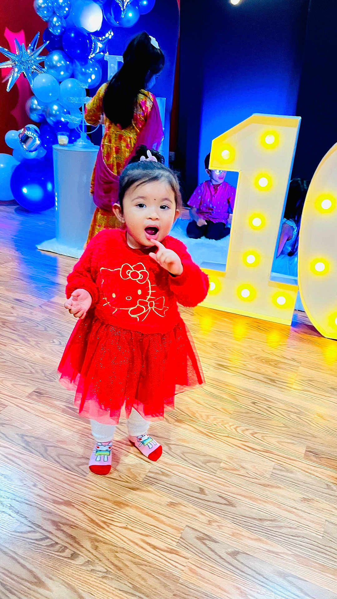 Aratrika joined the competition — help win amazing prizes! toddler, child, red_dress, hello_kitty, balloons, birthday, number_one, party, floor, wooden_floor, celebration, indoor, lighting, person, socks, cute, decorations, smiling, people, event