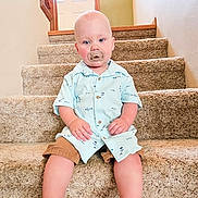 Cayden is registered to the contest to win money with this photo: toddler, child, pacifier, stairs, carpet, shirt, shorts, socks, indoor, door, railing, wall, baby, sitting, person, young_child, light, expression, calm, home