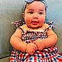 baby, blue_headband, checkered_dress, child, chubby_cheeks, clothing, couch, cute, hands, happy, headband, indoor, infant, person, portrait, red_white_blue, seated, smiling, soft_furniture, tongue_out