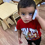 Damian Politz is registered to the contest to win money with this photo: toddler, child, boy, toy_story, red_shirt, phone, indoor, classroom, wooden_floor, small_chairs, table, looking_up, curious, expression, casual_clothing, playing, young_child, person, floor, furniture