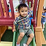 child, toddler, playground, slide, smiling, plaid_shirt, shorts, sneakers, outdoor, play_equipment, green_slide, artificial_grass, happy, cute, fun, kids, recreation, young_child, daylight, playing