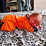 baby, bed, bedroom, blanket, cozy, drawer, face, furniture, hand, infant, lying_down, newborn, orange_outfit, patterned_blanket, person_partial, portrait, sleeping, smile, socks, soft_texture