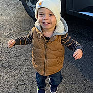 Patrick is registered to the contest to win money with this photo: asphalt, beanie, car, child, jacket, outdoor, pants, parking_lot, pavement, portrait, shadow, shoes, smiling, standing, sunlight, suv, toddler, vehicle, vest, winter_clothing