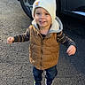 asphalt, beanie, car, child, jacket, outdoor, pants, parking_lot, pavement, portrait, shadow, shoes, smiling, standing, sunlight, suv, toddler, vehicle, vest, winter_clothing