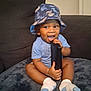 Tylan joined the competition — help win amazing prizes! toddler, child, hat, bucket_hat, sitting, smiling, happy, indoor, couch, blue_shirt, socks, baby, playful, face, person, furniture, soft_surface, chewing, black_object, cute