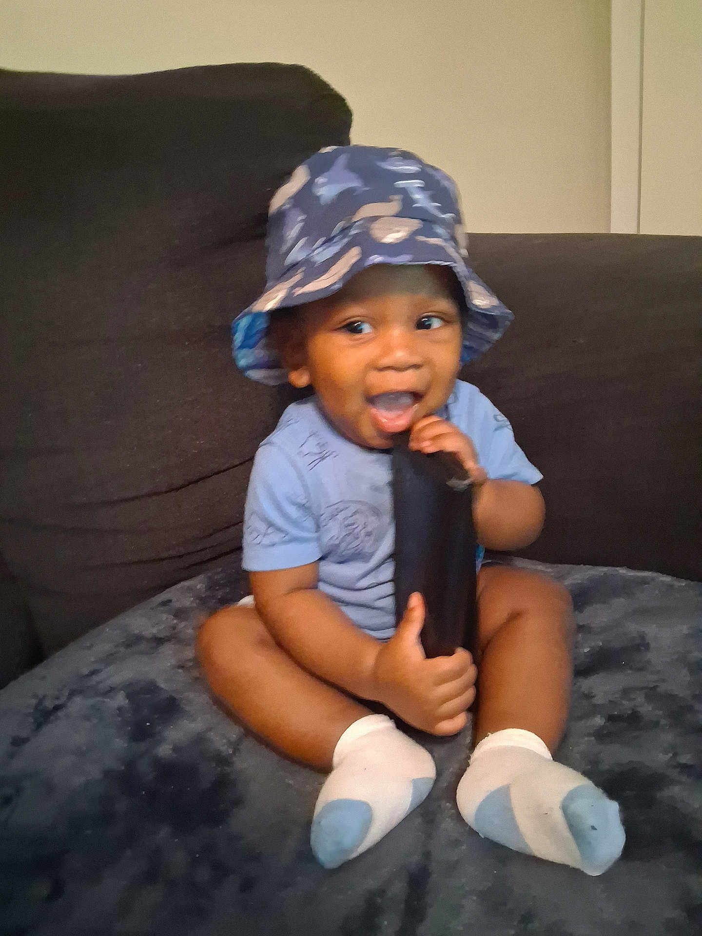 Tylan joined the competition — help win amazing prizes! toddler, child, hat, bucket_hat, sitting, smiling, happy, indoor, couch, blue_shirt, socks, baby, playful, face, person, furniture, soft_surface, chewing, black_object, cute