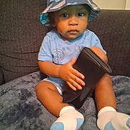 Tylan is registered to the contest to win money with this photo: toddler, child, hat, bucket_hat, blue_clothing, socks, wallet, indoor, sofa, blanket, seated, curious, person, face, hand, legs, cozy, cushion, home, casual