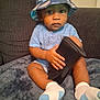 toddler, child, hat, bucket_hat, blue_clothing, socks, wallet, indoor, sofa, blanket, seated, curious, person, face, hand, legs, cozy, cushion, home, casual