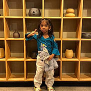 Sansa joined the competition — help win amazing prizes! child, girl, stuffed_animal, wolf, peace_sign, crocs, white_pants, teal_top, wooden_shelf, pottery, indoor, concrete_floor, standing, smiling, casual_clothing, cute, toy, person, young, decor