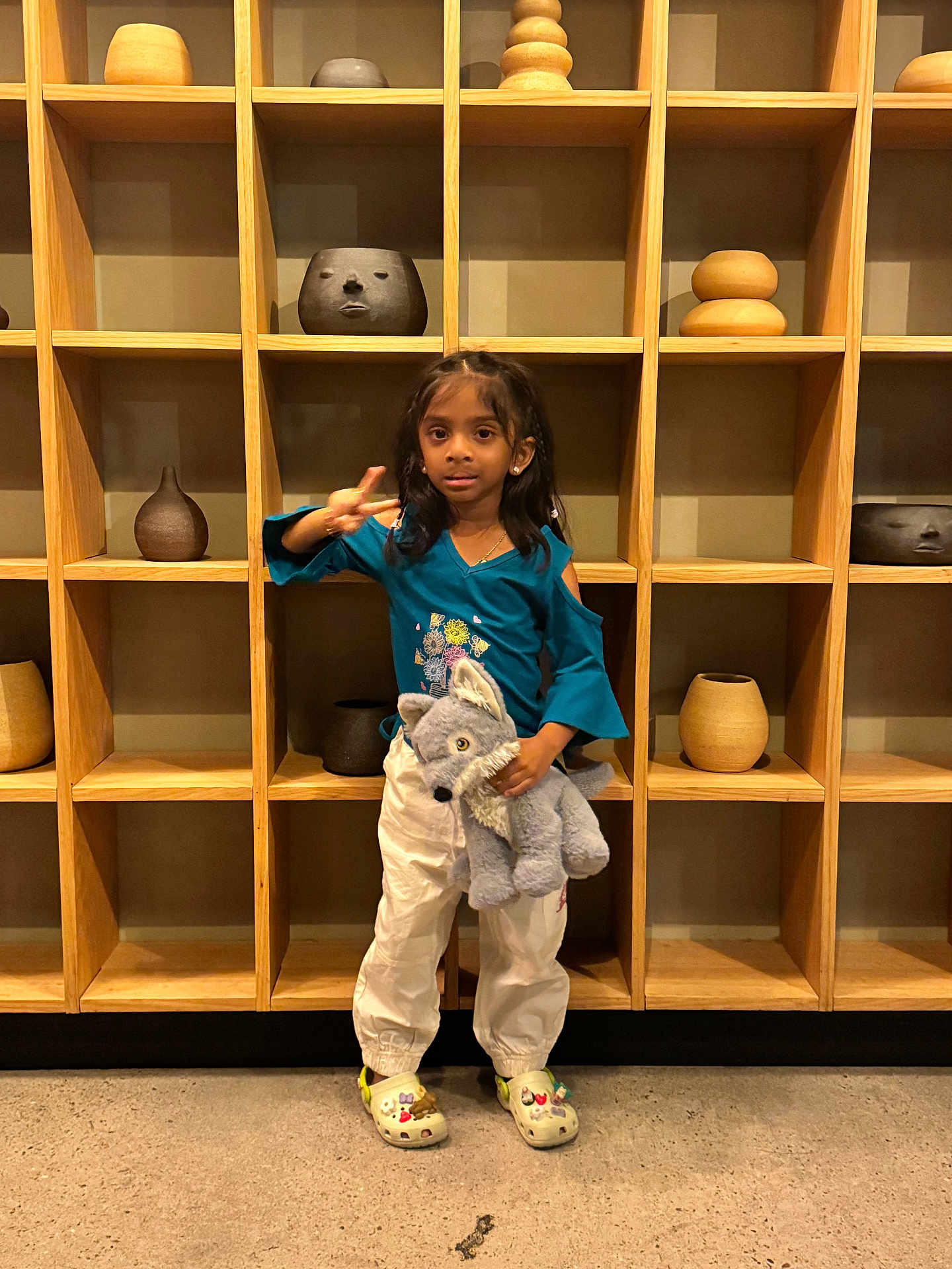 Sansa joined the competition — help win amazing prizes! child, girl, stuffed_animal, wolf, peace_sign, crocs, white_pants, teal_top, wooden_shelf, pottery, indoor, concrete_floor, standing, smiling, casual_clothing, cute, toy, person, young, decor