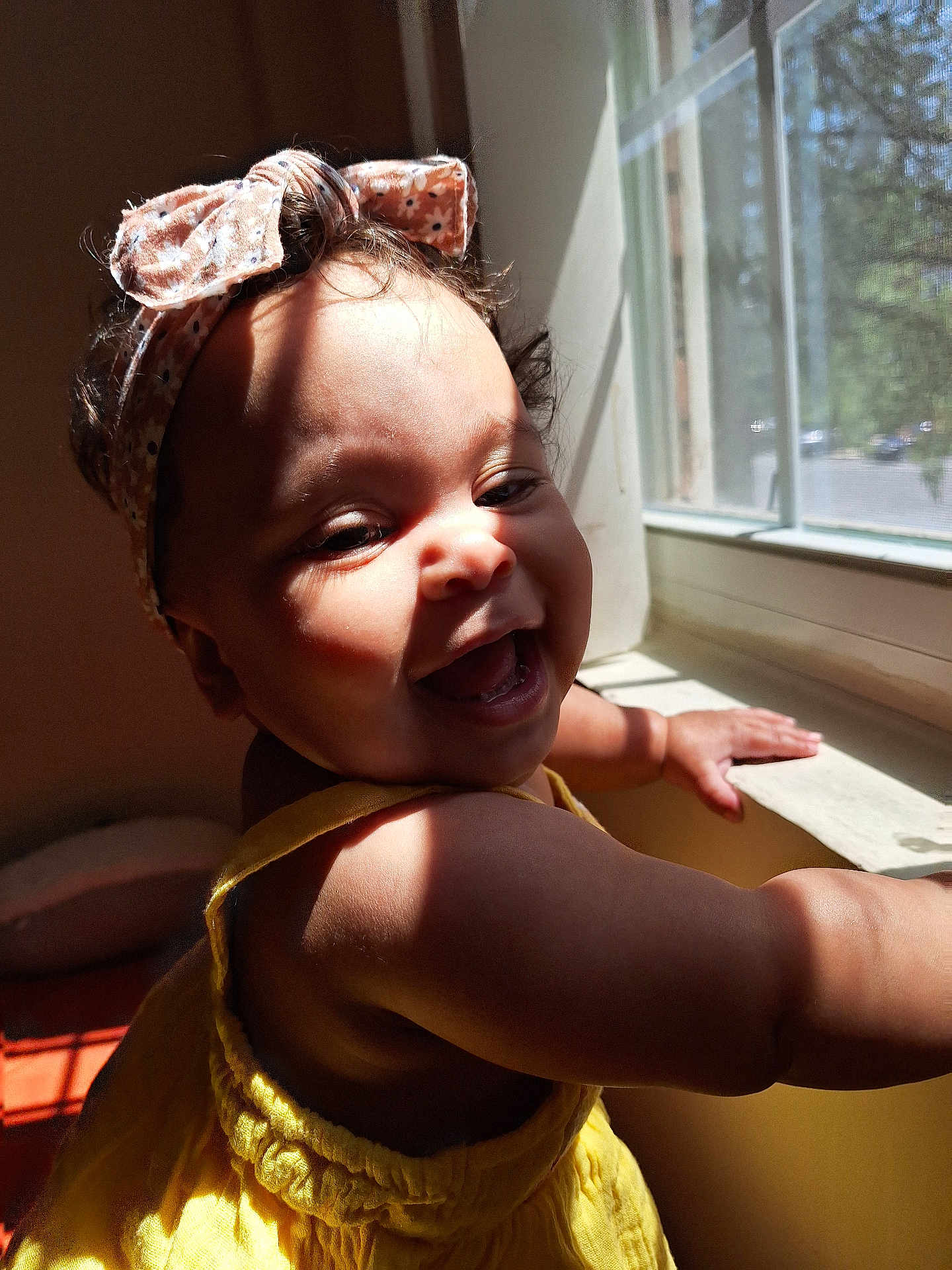 Victoria is registered to the contest to win money with this photo: baby, child, smiling, happy, window, sunlight, shadow, headband, yellow_dress, indoor, portrait, face, arm, skin, cute, infant, young_child, daylight, playing, expression