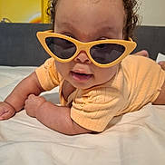 Victoria is registered to the contest to win money with this photo: baby, child, sunglasses, yellow, bed, cute, smiling, curly_hair, infant, clothing, indoor, portrait, lying_down, playful, person, face, fashion, accessory, eyewear, skin