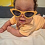 baby, child, sunglasses, yellow, bed, cute, smiling, curly_hair, infant, clothing, indoor, portrait, lying_down, playful, person, face, fashion, accessory, eyewear, skin