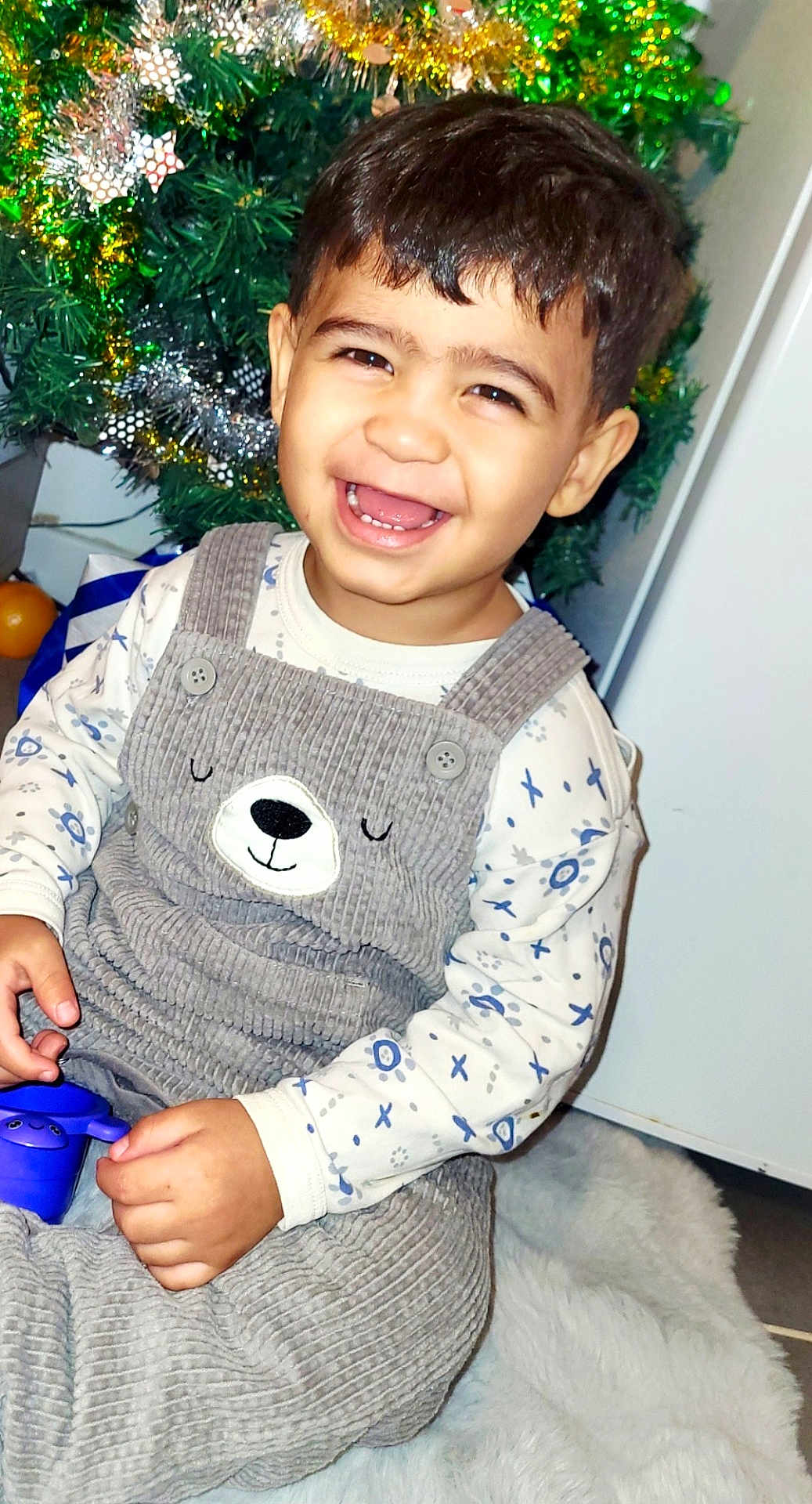 Alex a rejoint le concours — aidez-le/la à gagner de superbes lots ! toddler, child, smiling, overalls, bear_face, toy, purple_toy, sitting, fluffy_rug, christmas_tree, holiday_decorations, festive, indoors, happy, cute, clothing, face, person, celebration, joy