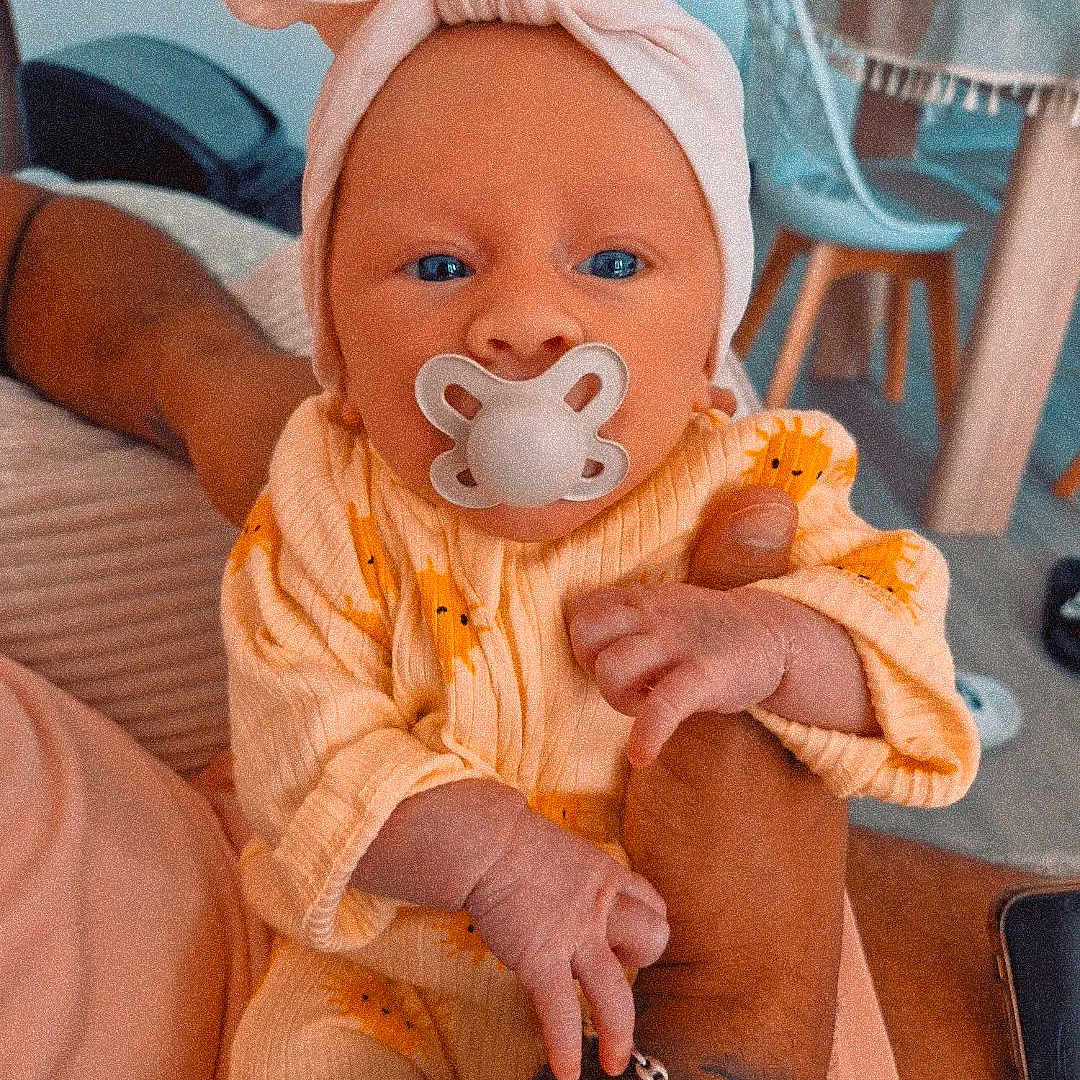 Vahina participe au concours pour gagner de l'argent avec cette photo : arm, baby, blue_eyes, chair, clothing, cute, furniture, hand, headwrap, holding, home, indoor, infant, pacifier, person, portrait, skin, sleepsuit, table, tattoo