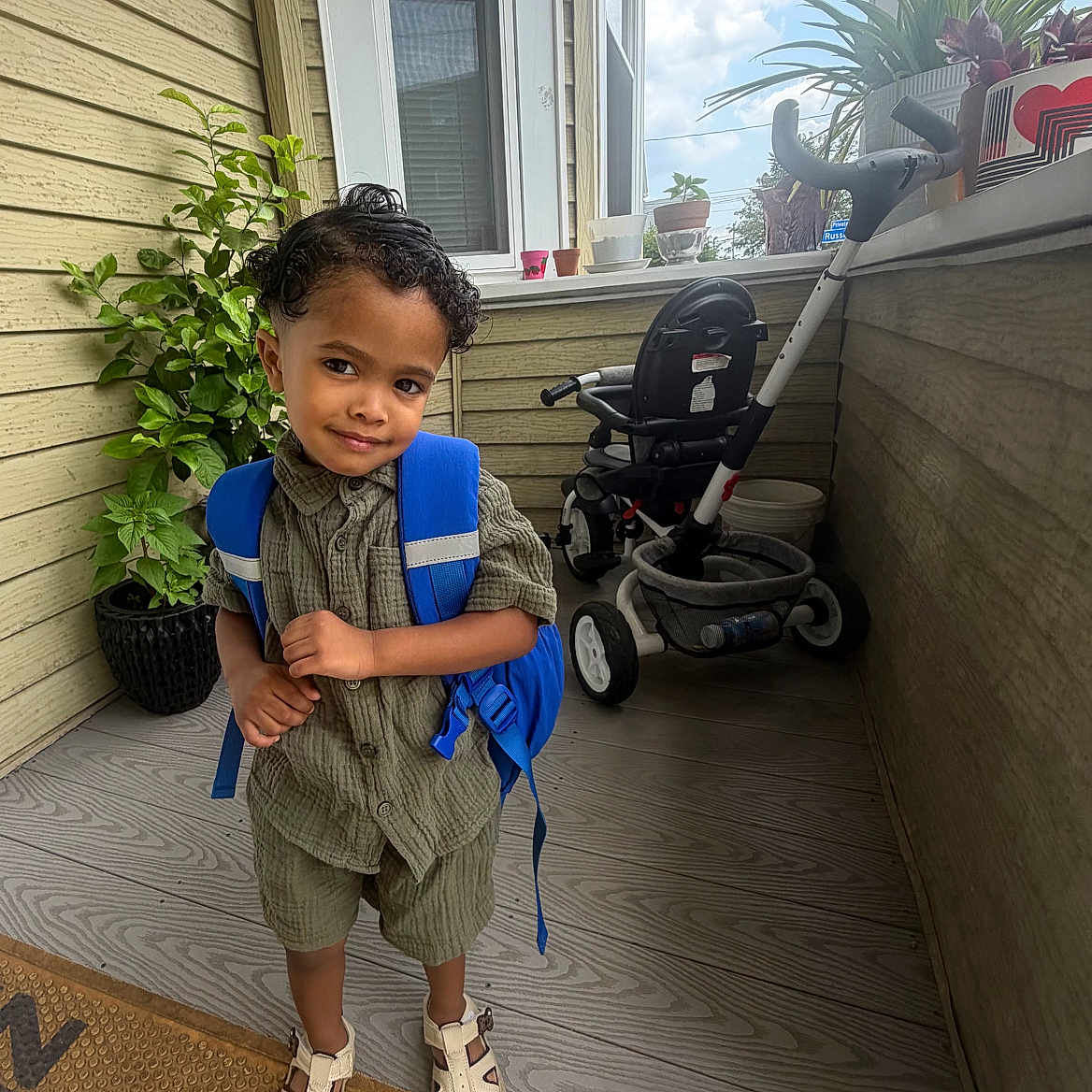 Gusttavo joined the competition — help win amazing prizes! child, toddler, portrait, standing, smiling, backpack, school_ready, outdoor, porch, house, plant, stroller, window, sunny, sky, wooden_floor, sandals, clothing, curly_hair, person