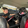 animal, backseat, baseball_bat, black_dog, boy, car_interior, casual_clothing, child, daytime, dog, happy, leather_seat, petting, playing, road, seatbelt, sunlight, tongue_out, travel, window