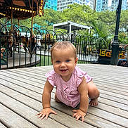 Leila joined the competition — help win amazing prizes! baby, child, crawling, pink_clothing, outdoor, wooden_floor, urban_park, carousel, fence, trees, buildings, daylight, happy, smiling, person, playground, nature, baby_girl, curious, cute