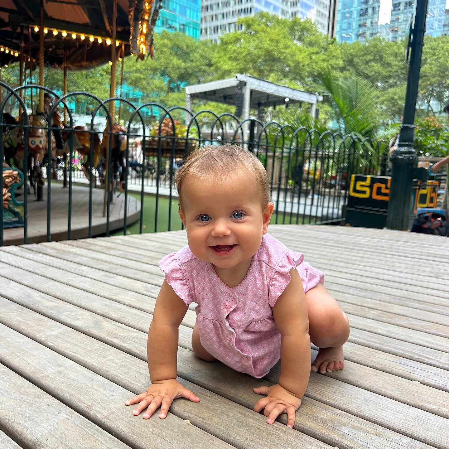 Leila joined the competition — help win amazing prizes! baby, baby_girl, buildings, carousel, child, crawling, curious, cute, daylight, fence, happy, nature, outdoor, person, pink_clothing, playground, smiling, trees, urban_park, wooden_floor