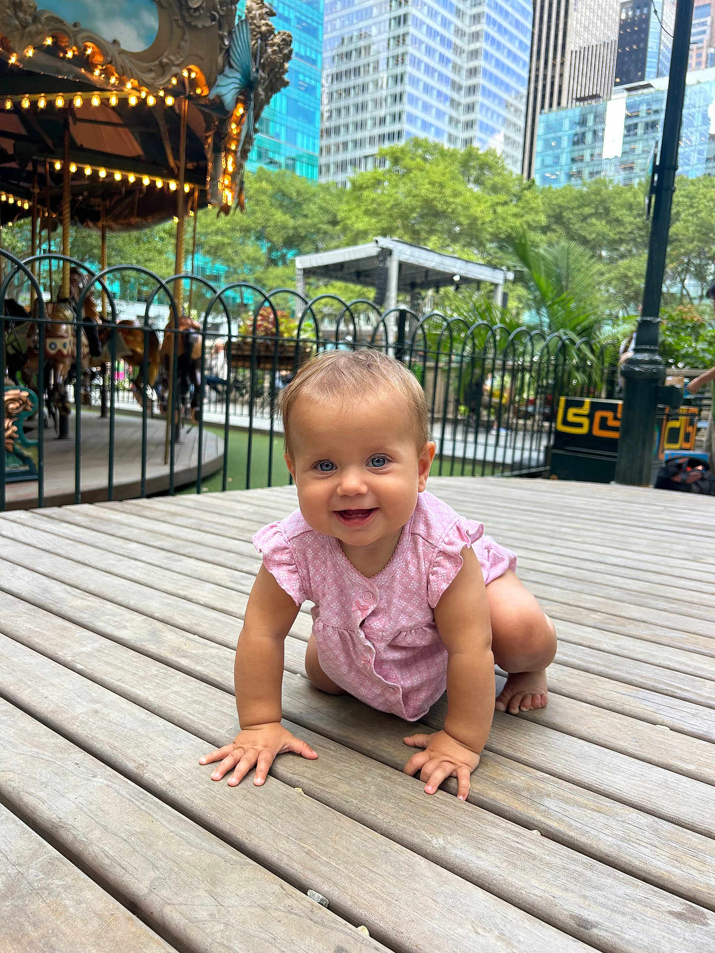 Leila joined the competition — help win amazing prizes! baby, child, crawling, pink_clothing, outdoor, wooden_floor, urban_park, carousel, fence, trees, buildings, daylight, happy, smiling, person, playground, nature, baby_girl, curious, cute