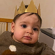 Zaïen participe au concours pour gagner de l'argent avec cette photo : baby, child, crown, golden_crown, sweater, dog_design, indoor, wooden_table, curious_expression, face, head, hand, clothing, portrait, person, furniture, warm_lighting, cozy, young_child, seated