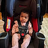 Laelani is registered to the contest to win money with this photo: baby, infant, child, car_seat, baby_car_seat, pink_clothing, straps, sitting, indoors, flooring, rug, black, red, serious_expression, person, cute, young_child, portrait, comfortable, safety