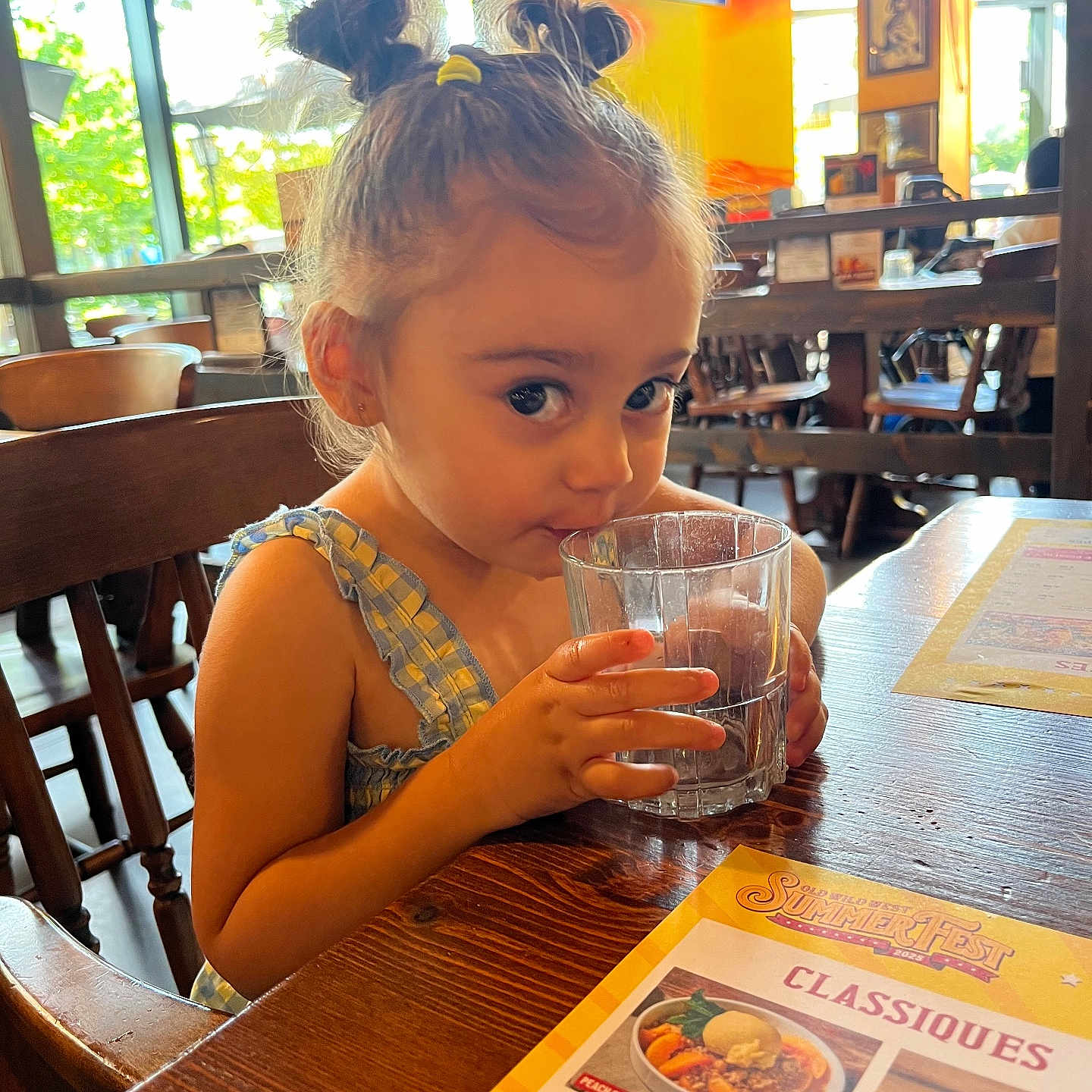 Eleysa a rejoint le concours — aidez-le/la à gagner de superbes lots ! casual_clothing, child, curious, cute, daylight, drinking, furniture, girl, glass, hair_buns, indoor, menu, menu_card, person, restaurant, seated, small_person, table, water, wooden_chair