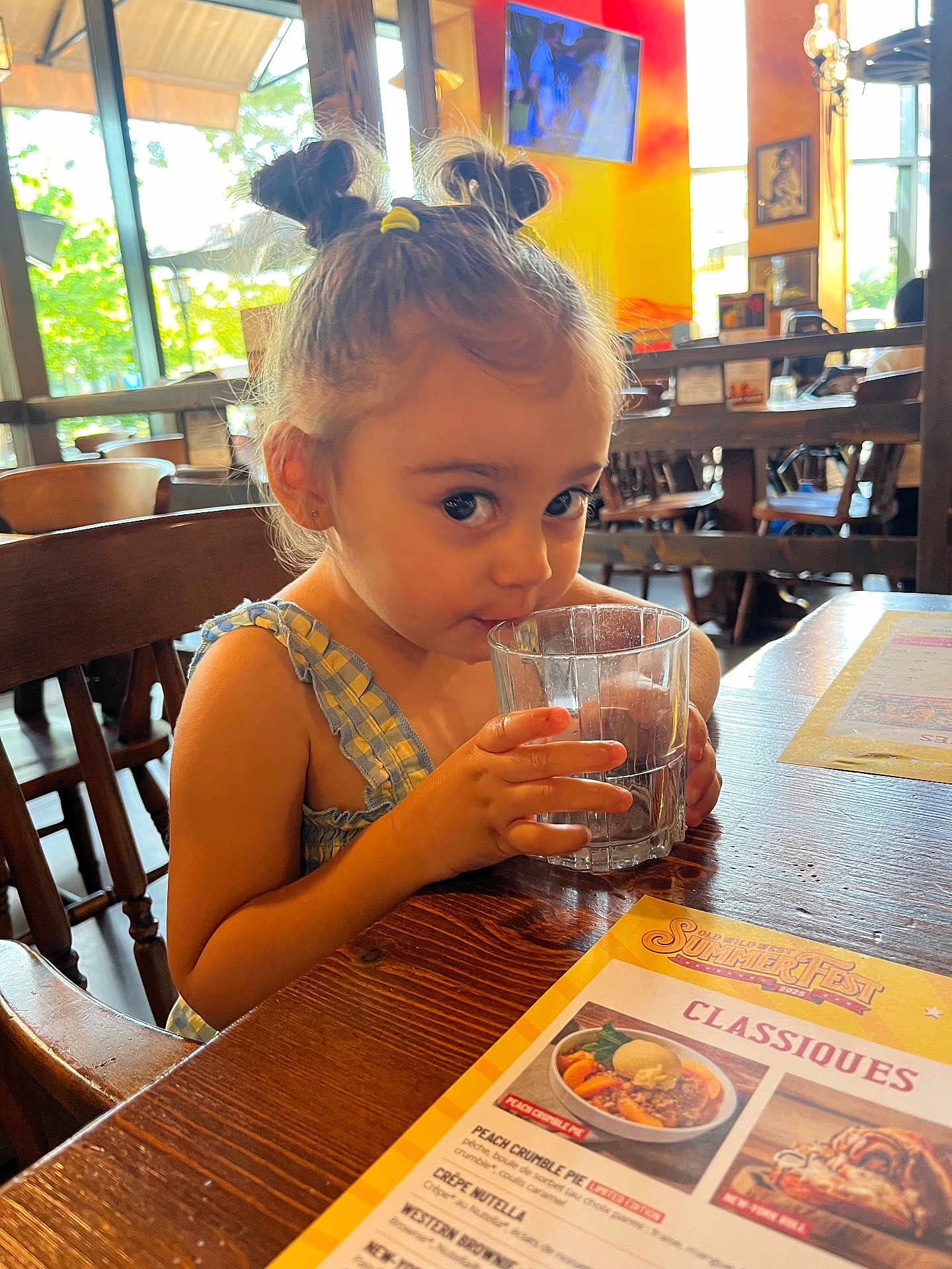 Eleysa a rejoint le concours — aidez-le/la à gagner de superbes lots ! child, girl, drinking, glass, water, restaurant, table, menu, wooden_chair, hair_buns, cute, indoor, daylight, curious, person, casual_clothing, small_person, seated, furniture, menu_card