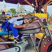 Alexa joined the competition — help win amazing prizes! child, toddler, carnival, amusement_ride, motorcycle_ride, blue_pants, white_shirt, fairground, colorful_lights, outdoor, daytime, playful, fun, seat, handlebars, young_child, casual_clothing, people, entertainment, smiling
