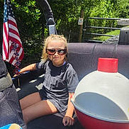 Alexa is registered to the contest to win money with this photo: child, sunglasses, car_seat, american_flag, outdoor, sunlight, greenery, toy, casual_clothing, summer, happy, smiling, person, seatbelt, vehicle, playful, red, white, blue, nature