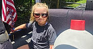 Alexa is registered to the contest to win money with this photo: american_flag, blue, car_seat, casual_clothing, child, greenery, happy, nature, outdoor, person, playful, red, seatbelt, smiling, summer, sunglasses, sunlight, toy, vehicle, white