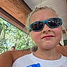 child, children, portrait, selfie, sunglasses, serious_expression, name_tag, outdoor, summer, trees, patio, seating, white_shirt, blonde_hair, face, photobomb, sunlight, wooden_structure, casual, photography