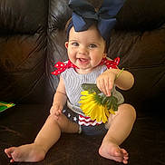 Aniyah is registered to the contest to win money with this photo: baby, child, smiling, sitting, flower, bow, headband, couch, leather, clothing, red, blue, yellow, happy, indoor, cute, portrait, skin, person, toddler