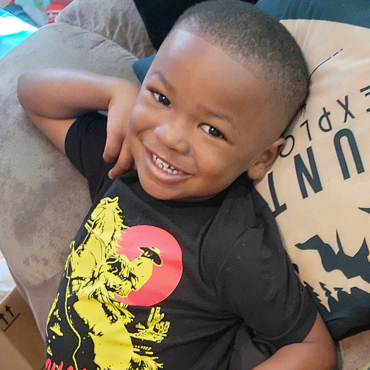 Maverick joined the competition — help win amazing prizes! child, smiling, portrait, indoors, couch, sitting, black_shirt, dinosaur_shirt, short_hair, happy, face, teeth, arm, hand, pillow, blanket, camouflage, home, casual, photography