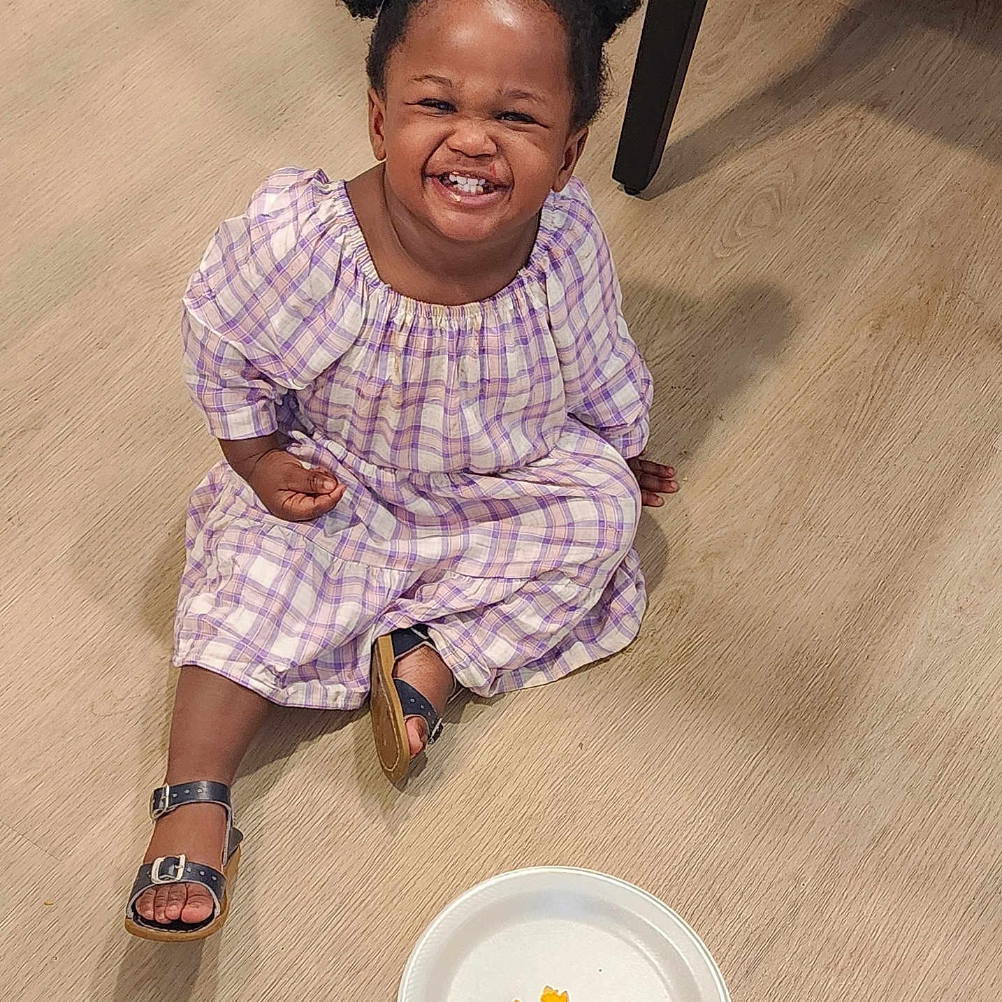 Miracle is registered to the contest to win money with this photo: toddler, child, smiling, happy, girl, pigtails, plaid_dress, sandals, floor, wooden_floor, plate, cheese_crackers, snack, indoor, person, cute, sitting, food, casual, portrait