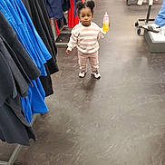 Miracle is registered to the contest to win money with this photo: bottle, casual_clothing, child, clothing_rack, clothing_store, curious, floor, flooring, hair_buns, indoor, pants, person, retail, shoes, shopping, sneakers, standing, sweatshirt, toddler, young_child