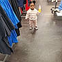 bottle, casual_clothing, child, clothing_rack, clothing_store, curious, floor, flooring, hair_buns, indoor, pants, person, retail, shoes, shopping, sneakers, standing, sweatshirt, toddler, young_child