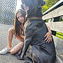 girl, dog, black_dog, pet, happy, smiling, bench, outdoor, sunlight, pavement, gate, greenery, leash, collar, casual_clothing, child, nature, friendship, crouching, playful