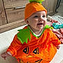baby, child, pumpkin_costume, orange, green, hat, smiling, indoor, floor, wooden_furniture, cute, happy, face, sitting, costume, holiday, festive, infant, clothing, playful