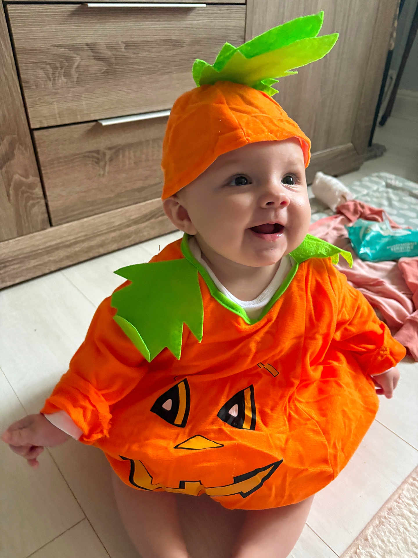 Ray-Anne is registered to the contest to win money with this photo: baby, child, pumpkin_costume, orange, green, hat, smiling, indoor, floor, wooden_furniture, cute, happy, face, sitting, costume, holiday, festive, infant, clothing, playful