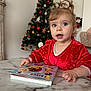 Lucie a rejoint le concours — aidez-le/la à gagner de superbes lots ! toddler, child, red_dress, blue_eyes, book, marble_table, christmas_tree, holiday_decor, indoor, curly_hair, festive, toy, face, person, home, furniture, smiling, cute, happy, decorations