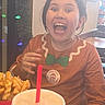 child, smiling, happy, indoor, restaurant, food, drink, waffle_fries, table, clothing, face, person, holiday_lights, decorations, brown_hair, long_sleeve_shirt, gesture, mouth_open, fun, casual
