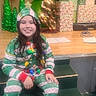 Mateo is registered to the contest to win money with this photo: child, christmas_pajamas, holiday_lights, green_steps, wrapped_presents, christmas_trees, small_tree, stage, festive, smiling, indoor, holiday_decor, long_hair, seated, festive_hat, floor, wooden_floor, green, happy, person